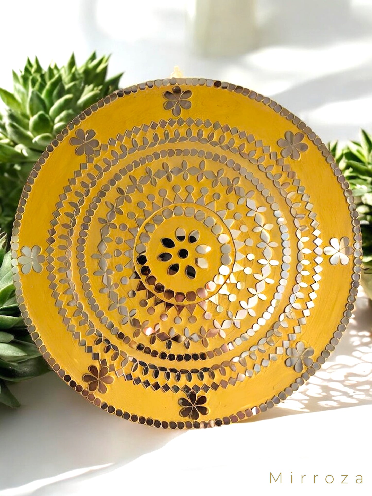 Close-up of handmade yellow round floral wall art with mosaic detailing, hanging on white wall with sunlight and flowers